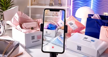 Bright glossy magazine-style image showing a smartphone filming a modern unboxing scene with ecommerce parcels, shopping bags, tissue paper, and luxury accessories, hinting at AI-driven returns fraud through subtle visual mismatches between the real setup and the phone screen. 